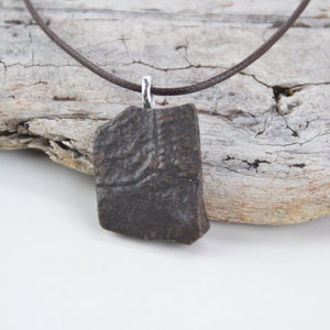 May include: A brown ceramic pendant with a textured surface hangs from a brown leather cord. The pendant is attached to the cord with a silver loop.