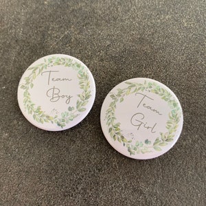 Peut inclure: Deux boutons ronds blancs avec des couronnes de feuilles vertes autour des bords. Le texte "Team Boy" est sur un bouton et "Team Girl" sur l'autre.