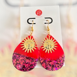 May include: A pair of teardrop-shaped earrings with a red and glittery pink design. Each earring features a gold sun face charm. The earrings are displayed on a white card with the text "KB Créations".