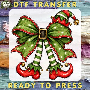 May include: A DTF transfer featuring a green polka-dot bow with a central black belt buckle, attached to striped green and white legs ending in curled red elf shoes with gold bells. The text reads DTF TRANSFER READY TO PRESS.