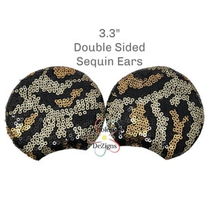 Peut inclure: Deux oreilles à sequins double face, chacune mesurant 8,4 cm, présentent un motif léopard noir et or. Les oreilles sont rondes et ont une base incurvée pour un port confortable. Le texte "Double Sided Sequin Ears" est affiché au-dessus des oreilles.