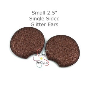 Peut inclure: Deux petits accessoires en forme d'oreille, marron et pailletés. Le texte sur l'image indique "Small 2.5" Single Sided Glitter Ears". Les accessoires sont probablement destinés à l'artisanat ou à la conception de costumes.