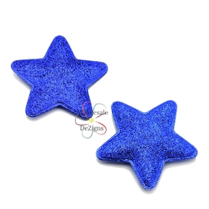 May include: Two blue glitter star-shaped embellishments. The stars have a five-point design and a textured, sparkling surface. The image is set against a white background. The text "Wholesale DeZigns" is in the lower left corner.