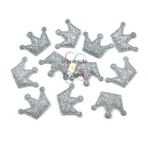 Puede incluir: Una colección de adornos de corona de purpurina plateada sobre un fondo blanco. Las coronas son pequeñas, con un diseño real clásico. La purpurina añade un efecto brillante, ideal para manualidades y decoraciones.