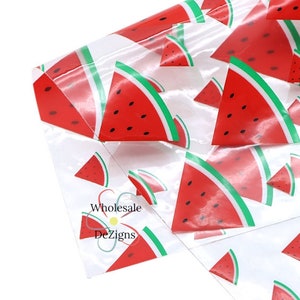 May include: Clear cellophane packaging featuring a repeating pattern of watermelon slices. The watermelons are red with black seeds and green rinds. The text "Wholesale Dezigns" is printed on the packaging.