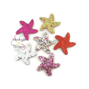 May include: A collection of six glittery starfish in various colors, including pink, gold, red, silver, and a multicolored confetti design. The starfish are arranged on a white background. The text "Wholesale Dezigns" is in the image.