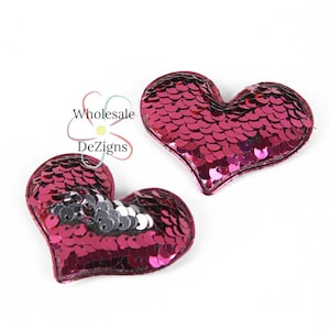 May include: Two heart-shaped patches covered in shiny, magenta sequins. The patches have a dark pink border and are set against a white background. The patches are slightly different sizes. The text "Wholesale DeZigns" is in the upper left corner.