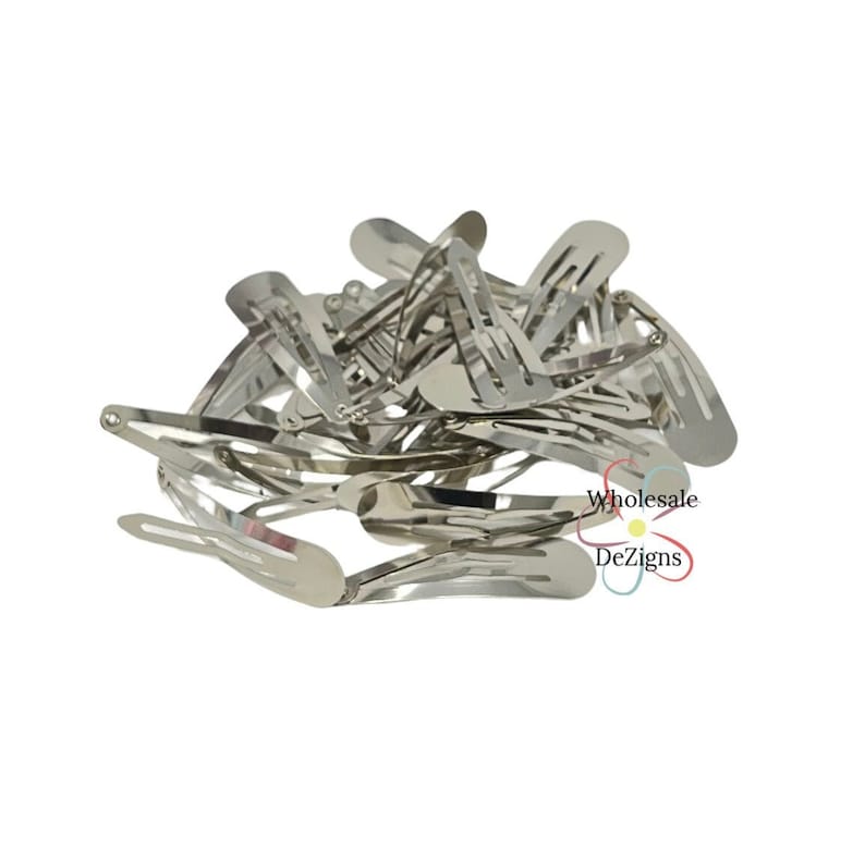 May include: A collection of silver-toned metal snap hair clips piled together in a cluster. Each clip features a classic oval shape with a hinged mechanism. The text Wholesale DeZigns is printed in black and red near the bottom right corner.