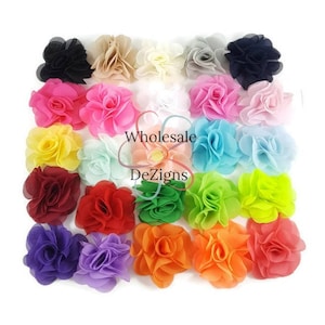 May include: A collection of twenty-five fabric flower appliques arranged in a grid, featuring a diverse spectrum of colors including black, beige, white, gray, pink, yellow, blue, red, green, purple, and orange. The text Wholesale DeZigns is visible.