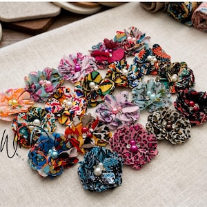 May include: A collection of fabric flower embellishments featuring diverse patterns including leopard print, floral, and geometric designs. Each flower is accented with a central bead or pearl. The text Wholesale is visible on the left side.
