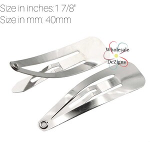 May include: Two silver-colored metal snap hair clips with a smooth, reflective finish. The clips measure 1 7/8 inches in length. The text "Wholesale DeZigns" is printed near the clips, accompanied by a small, colorful flower-shaped logo.