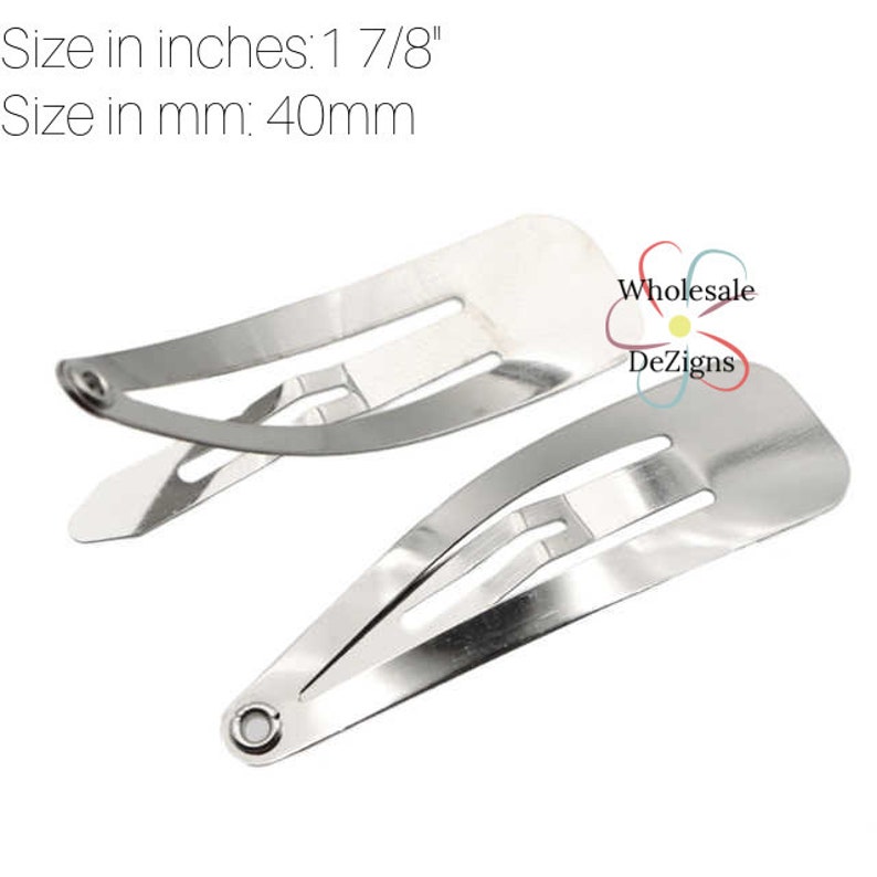 May include: Two silver-colored metal snap hair clips with a smooth, reflective finish. The clips measure 1 7/8 inches in length. The text "Wholesale DeZigns" is printed near the clips, accompanied by a small, colorful flower-shaped logo.