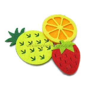 May include: Three felt fruit shapes including a yellow pineapple with green leaves, a bright orange citrus slice with yellow segments, and a red strawberry with a green top. The text wholesaledesigns.com is printed across the centre of the shapes.