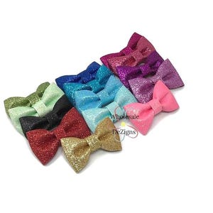 May include: A collection of glittery bow hair accessories in various colors, including pink, red, gold, black, green, blue, and purple. The bows are arranged in a diagonal row, showcasing their sparkly texture and design.