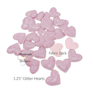 May include: A collection of pink glitter hearts, each measuring 1.25 inches. The hearts are scattered across a white surface, with some showing a fabric backing. The text "Wholesale DeZigns" and "Fabric back" are also visible.