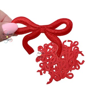 May include: Red embroidered bow patches. One patch is held by a hand, with a pile of additional patches in the background. The patches are a vibrant red color and feature a detailed bow design. The text "Wholesale Dezigns" is visible.