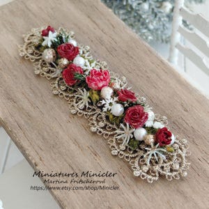 Könnte beinhalten: Eine dekorative Miniatur-Spitzenborte mit goldener Metallic-Optik, verziert mit kleinen roten Rosen, weißen perlenartigen Kugeln und winzigen weißen Schneeflocken-Akzenten. Das rechteckige Stück zeigt detaillierte florale und Spitzenmuster.