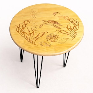 May include: Round wooden coffee table with a sea turtle and underwater scene etched into the light wood surface. The table has black hairpin legs. The design includes seaweed, bubbles, coral, and seashells.