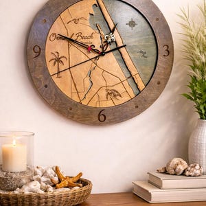 May include: A round wooden wall clock featuring a map of Ormond Beach, Florida. The clock has a dark brown frame and a light brown map design with blue accents representing the ocean. The clock hands are black and red.