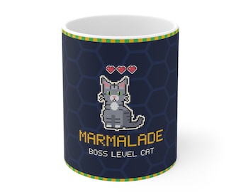 Puede incluir: Una taza de cerámica azul oscuro con un diseño de arte de píxeles de un gato atigrado gris con tres corazones rojos encima. El texto MARMALADE BOSS LEVEL CAT está impreso en una fuente de píxeles amarilla. La taza tiene un patrón de panal y un borde verde y amarillo.
