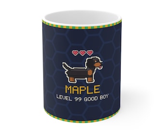 Puede incluir: Una taza de cerámica azul oscuro con un perro salchicha en pixel art, tres corazones rosas y el texto amarillo "MAPLE" sobre el texto blanco "LEVEL 99 GOOD BOY". La taza tiene un patrón hexagonal azul oscuro y un borde verde y amarillo.
