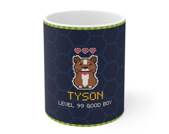 Puede incluir: Una taza de cerámica azul oscuro con un gráfico de pixel art de un perro marrón y blanco con tres corazones rojos encima. El texto TYSON y LEVEL 99 GOOD BOY está impreso en fuente pixelada amarilla y blanca sobre un patrón de panal.