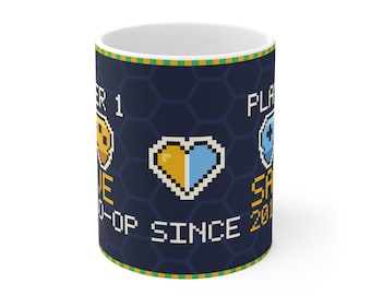 Puede incluir: Una taza de cerámica blanca con un patrón de fondo hexagonal azul oscuro. El diseño presenta gráficos pixelados que incluyen un mando de juego amarillo, uno azul y un corazón dividido en amarillo y azul. El texto dice PLAYER 1, PLAYER 2, CO-OP y SINCE 2018.