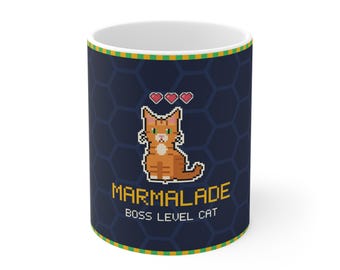 Puede incluir: Una taza de cerámica azul oscuro con un patrón hexagonal repetitivo. El frente presenta un gato atigrado naranja en pixel art con tres corazones rojos encima, el texto MARMALADE y el texto BOSS LEVEL CAT en fuente amarilla pixelada.