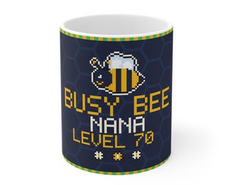 Puede incluir: Una taza de cerámica blanca con fondo azul oscuro y un patrón de panal. El diseño incluye un gráfico de abeja en estilo píxel y el texto amarillo BUSY BEE NANA LEVEL 70, decorado con tres pequeñas flores también en estilo píxel.