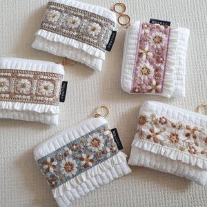 May include: Five white quilted pouches with gold-tone key rings. Each pouch features a central band of floral embroidery in various colors including blue, pink, and brown, accented with white ruffles and a small black tag with Korean text.