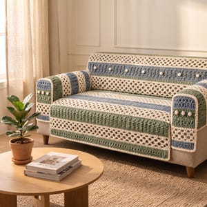 May include: A crochet sofa cover featuring horizontal stripes in cream, sage green, and dusty blue. The textile incorporates an openwork diamond pattern, textured ribbed sections, and small white pom-poms arranged in a linear row.