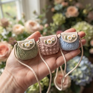 May include: Three miniature crochet bags in sage green, dusty pink, and light blue. Each bag features a cream-colored flap decorated with a white daisy embroidery, a gold-tone metal clasp, and a long, thin, braided cream-colored shoulder strap.