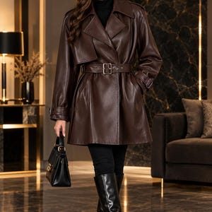 Peut inclure: Un trench-coat en cuir marron foncé doté d'un large col, de manches longues avec poignets à boucle et d'une ceinture assortie avec une boucle argentée. Le manteau est porté sur un col roulé noir, un pantalon noir et des bottes hautes noires.