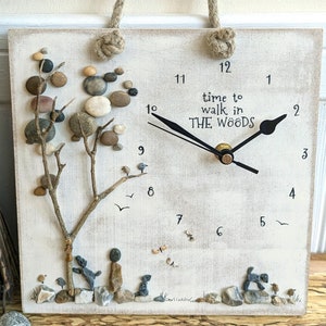 Puede incluir: Reloj cuadrado de madera con asa de cuerda. La esfera es blanquecina con números y manecillas negras. A la izquierda hay un árbol hecho de ramitas y guijarros. El texto "time to walk in the woods" está en el centro.