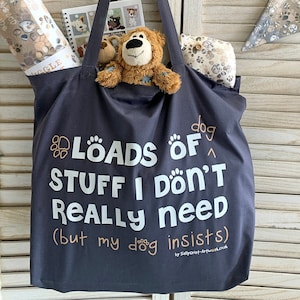 Peut inclure: Un sac fourre-tout bleu marine avec l'inscription "LOADS OF STUFF I DON'T REALLY NEED (but my dog insists)". Il contient un jouet en peluche, un carnet et des rouleaux de tissu à motifs de pattes de chien. Une bannière est visible.