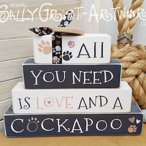 Puede incluir: Una pila de cuatro bloques de madera con las palabras "ALL YOU NEED IS LOVE AND A COCKAPOO" en letras blancas. Los bloques están pintados en blanco y azul marino, con huellas de patas y un diseño de corazón. Un pequeño adorno de madera con un diseño de huella de pata se encuentra encima.