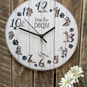 Könnte beinhalten: Eine runde Wanduhr mit hellem Holzmaserungshintergrund. Das Zifferblatt zeigt Illustrationen von Hunden, Pfotenabdrücken und den Schriftzug "time for DOGS!". Schwarze Zeiger zeigen die Uhrzeit an. Die Uhr ist an einer Holzwand befestigt.
