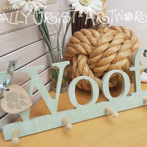 May include: A light green wooden wall hook with the word "Woof" cut out. The hook has three pegs and a heart-shaped tag with a paw print design. A ribbon with paw prints is attached to the tag. The background includes a decorative rope knot and flowers.