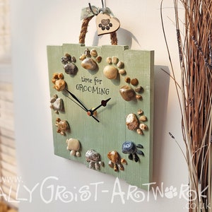 Op de afbeelding: Een lichtgroene houten klok met een touw handvat en een pootafdruk bedel. De klok heeft pootafdrukken van stenen en de tekst "time for GROOMING". De wijzers zijn zwart.