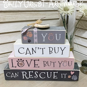 Puede incluir: Una pila de cuatro bloques de madera con las frases "YOU", "CAN'T BUY", "LOVE BUT YOU" y "CAN RESCUE IT" en letras blancas y rosas. Los bloques son grises, blancos y rosas con huellas de patas y una cinta decorativa.