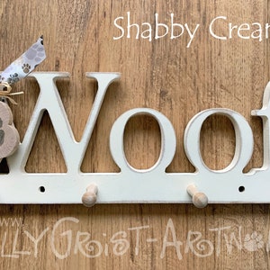 May include: A cream-colored wooden wall-mounted dog leash holder with the word "Woof" in large letters. It has three wooden pegs for hanging leashes and a paw print-shaped tag. The text "Shabby Cream" is above the word "Woof".