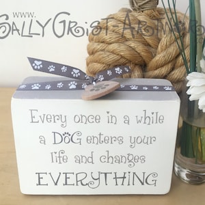 May include: A white wooden block with a grey top, inscribed with the words "Every once in a while a DOG enters your life and changes EVERYTHING." It's decorated with a grey paw print ribbon and a wooden heart charm. A charming gift for dog owners.
