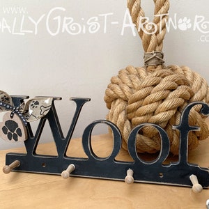 May include: Black wooden "Woof" wall hook with three pegs, a paw print tag, and a decorative bow. The background features a large rope knot. The text "www. Sally Groist-ArtWork.co.uk" is visible.