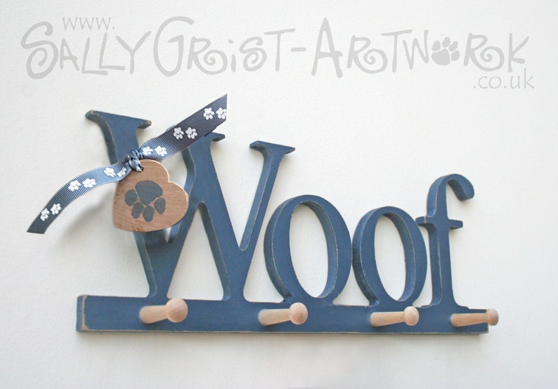 May include: A navy blue wooden wall-mounted coat rack spelling "Woof" with three pegs. A wooden heart-shaped tag with a paw print hangs from a blue and white paw print ribbon. The sign is for pet owners.