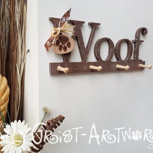Op de afbeelding: Een bruine, houten wandkapstok met het woord "Woof". Het heeft vier houten pinnen om items op te hangen en een hartvormig label met een pootafdruk. Een decoratieve strik en raffia accentueren de bovenkant.