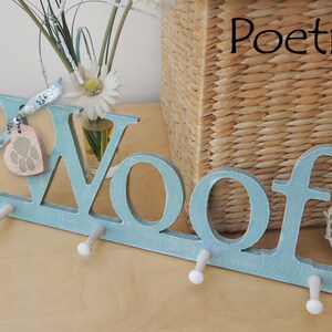May include: A light blue wooden wall hook with the word "Woof" in large letters. The hook has three pegs and a paw print charm. The word "Poetic" is written in black script in the upper right corner. A woven basket and flowers are in the background.