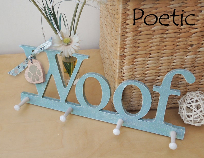 May include: A light blue wooden wall hook with the word "Woof" in large letters. The hook has three pegs and a paw print charm. The word "Poetic" is written in black script in the upper right corner. A woven basket and flowers are in the background.