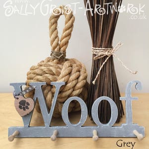 May include: A grey wooden "Woof" key holder with three pegs, a heart-shaped tag with a paw print, and a decorative rope knot. The word "Grey" is written in the bottom right corner. A bundle of dried sticks is in the background.