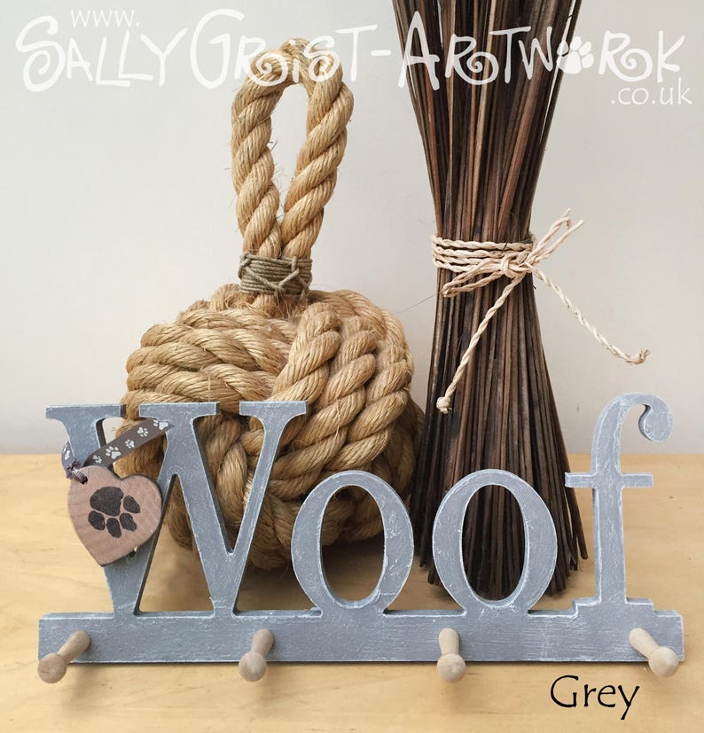 May include: A grey wooden "Woof" key holder with three pegs, a heart-shaped tag with a paw print, and a decorative rope knot. The word "Grey" is written in the bottom right corner. A bundle of dried sticks is in the background.