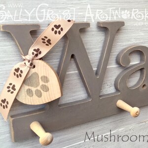 May include: A gray wooden wall hook with the letters "VA" and a small "a". It has two wooden pegs and a wooden tag with a paw print, tied with a ribbon printed with paw prints. The word "Mushroom" is at the bottom.